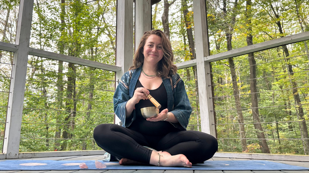Eve Efron is a Yoga Teacher seated on a blue yoga mat in a the corner of a room with tall, screened windows looking over lush, green trees in the woods. Eve is a white woman with dark blonde hair, wearing black leggings, a blue jacket and holding a brass singing bowl and wooden mallet.